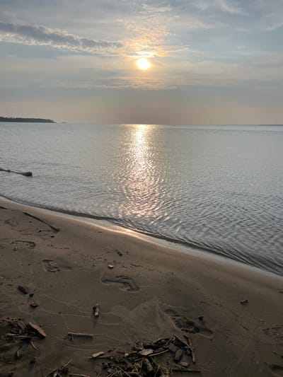 The image depicts a serene natural scene at what appears to be a beach or shoreline. The sun is low in the sky, casting a warm glow and reflecting off the gentle ripples of the water. The sky is partly cloudy, with the sun partially obscured by clouds. In the distance, a treeline is visible along the horizon. The foreground is a sandy beach littered with small pieces of driftwood and scattered debris. Footprints are visible in the sand, indicating recent human presence. The overall impression is one of tranquility and natural beauty. There is no indication of the city of Borovoye, Russia within the image.