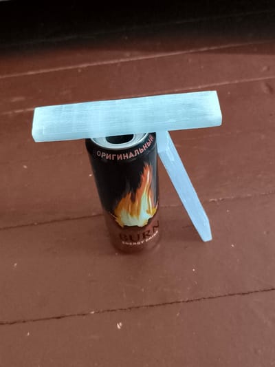A light blue, translucent selenite crystal is placed horizontally across the top opening of a black energy drink can. The can has a flame design and text that reads "ORIGINAL" in Russian and "BURN ENERGY DRINK" in English. A second, smaller piece of the same selenite crystal leans against the can, extending downwards. The objects are positioned on a dark brown, wooden surface with visible plank lines and some light-colored debris. The image is a close-up shot with shallow depth of field, focusing on the selenite and the can. There is no indication of location within the city of Yasynivka, Ukraine.