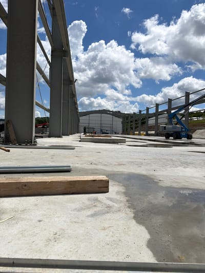 The image depicts an outdoor construction site, likely in Skrzydlna, Poland, under a partly cloudy sky. The foreground shows a concrete surface with puddles of water and a wooden beam. In the midground, a large, unfinished steel structure is visible, with prominent gray support columns and beams. A blue boom lift, identified as a Genie S-85, is parked on the concrete. In the background, another section of the steel structure is under construction, with white siding on a building. Construction machinery, including a red excavator, is present further in the background. The environment appears to be a rural or semi-rural area with a wooded hillside visible in the distance.