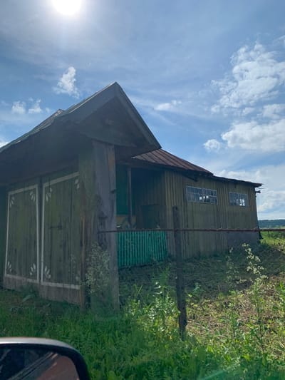 A weathered, green wooden structure with a rusty metal roof stands in a rural outdoor setting. The building has a pitched roof and appears to be a barn or storage shed. It features two large, double doors on the front, each adorned with white painted leaf motifs. To the right of the doors, a section of the building has several windows, appearing to be divided into panes, and a green corrugated metal fence is visible in front of this section. The structure is surrounded by lush green grass and vegetation, with a wire fence running along the front. In the background, rolling hills are visible under a bright blue sky with scattered white clouds and a bright sun. The image was captured from a low angle, possibly from within a vehicle, as evidenced by a portion of a car's side mirror in the bottom left corner. The location is identified as Barsai, Russia. There are no people or animals visible in the image.
