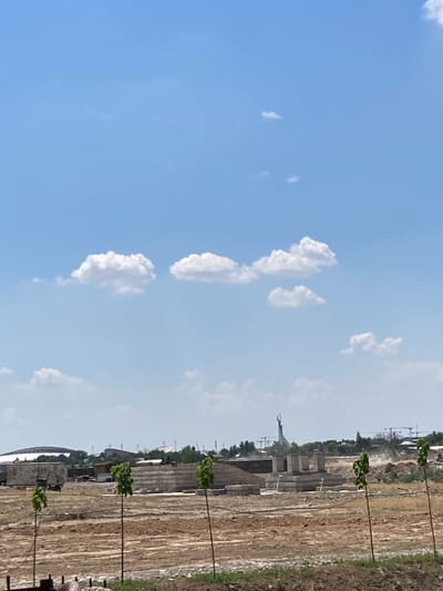 The image depicts a construction site under a clear blue sky with scattered white clouds. In the foreground, there are three young trees planted in a line, each supported by a thin pole. The ground is dry and uneven, with visible soil and some debris. In the middle ground, a partially constructed building made of concrete blocks is visible, along with piles of construction materials. Further in the background, several cranes suggest ongoing construction activities. On the left side of the background, a large, domed stadium structure can be seen. Dominating the far background is a distinctive tall monument with a circular element at its apex, a landmark in the city of Akbarak, Uzbekistan. The overall scene conveys a sense of development and urban expansion.