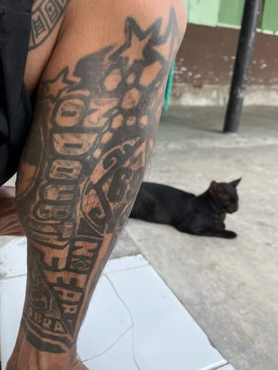 The image shows a close-up of a person's tattooed arm, with a black cat lying down in the background. The tattoo is intricate and covers a significant portion of the arm. The cat is lying on a concrete surface, and the background appears to be an outdoor setting with a wall and a pole. The overall impression is a candid, everyday moment captured in a somewhat urban or semi-urban environment.
In Tambakreja, Indonesia, this scene could depict a common sight in a residential area. The tattooed arm suggests a local individual, possibly an adult. The black cat is a domestic animal often found in Indonesian households and public spaces. The concrete floor and the wall could be part of a house or yard, or even a small business. The presence of a tattoo, while not exclusive to any particular social group, can be seen in various communities across Indonesia, including Tambakreja. The image evokes a sense of casual life, with the person relaxing while their pet rests nearby, a scenario that is easily imaginable within the context of Tambakreja's social fabric.
