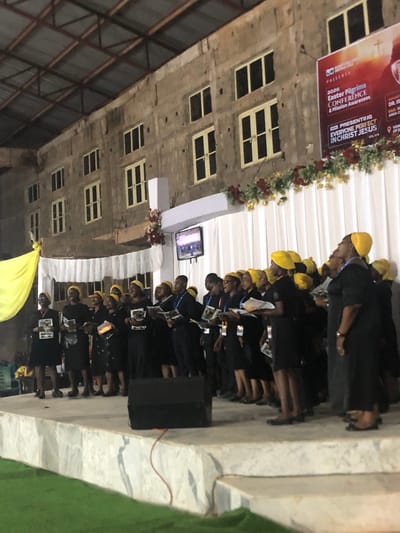 A photo taken in Nteje, Nigeria, displays a group of people, primarily wearing yellow headwear and dark attire, standing on a stage. A banner indicating the "2026 Easter Pilgrims Conference & Mission Awareness" event is visible. Buildings with multiple windows and a speaker are also visible in the background. The individuals appear to be engaged in some form of activity, potentially a religious service or conference event.