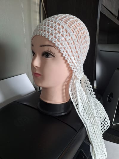 The image shows a white crocheted head covering on a mannequin head, placed on top of a black printer. The mannequin head has realistic features, including painted eyelashes and lips. The crocheted head covering is open-weave and has a decorative fringe. The background is blurred, but appears to be an indoor setting with shelves and monitors. There is no information in the image to determine the location in Fort Walton Beach, United States of America.