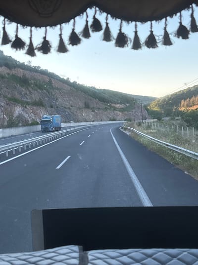 The image depicts a view from inside a vehicle looking out at a highway. A blue semi-trailer truck is traveling on the highway in the left lane. The truck has a yellow trailer with blue lettering. The highway is flanked by a concrete barrier and a metal guardrail on the left, and a grassy area with a fence on the right. The landscape beyond the highway consists of a steep, rocky hillside on the left and rolling, tree-covered hills on the right. The sky is clear and bright, suggesting it is daytime. The scene is set in or near Avra, Greece.
