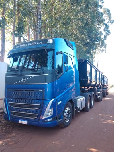 A blue Volvo FH truck with a "Globetrotter" designation is parked on a dirt road in São Gabriel do Oeste, Brazil. The truck is a tractor-trailer with a dark-colored Randon trailer attached, which appears to be a multi-axle dump trailer. The truck has a license plate that reads "SME9J11." The scene is illuminated by sunlight, and trees with dense foliage are visible in the background. A utility pole with wires runs vertically behind the truck. The ground is composed of reddish-brown dirt, with some fallen leaves scattered on it.