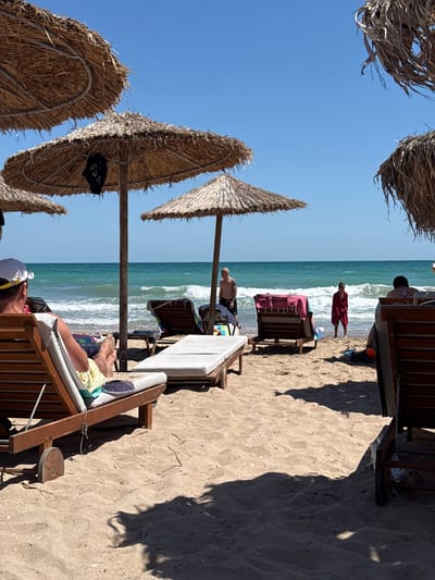 The image depicts a sunny day at the beach in Shabla, Bulgaria. Several thatched umbrellas provide shade over wooden lounge chairs arranged on the sand. The turquoise sea with gentle waves is visible in the background, with a clear blue sky above.
In the foreground, a person wearing a white cap and yellow and green patterned shorts is seated in a lounge chair, facing away from the camera and towards the sea. Their legs are extended, and their feet are visible. To the right of this person, another empty lounge chair with a white cushion is placed.
Further back, near the water's edge, a man with tattoos is standing, looking towards the sea. A person wrapped in a red towel is standing in the shallow water, facing the land. Other lounge chairs and umbrellas are visible in the middle ground and background. The sand is light brown and appears to be a typical beach texture, with some scattered debris. Shadows from the umbrellas and possibly vegetation are cast on the sand.