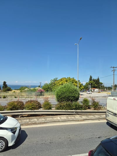 The image shows a bright, sunny day in Pylaia Municipal Unit, Greece. The foreground features a road with two vehicles, a white car on the left and a white van partially visible on the right. A metal guardrail separates the road from a median strip with various bushes and small trees. Beyond the median, there is a wooden fence and more greenery, including a large tree with yellow blossoms and a vibrant pink flowering bush. Further in the distance, the blue sea is visible under a clear blue sky, with a faint horizon line. A tall street lamp stands on the right side of the image, and power lines and a utility pole are also present in the background. The scene appears to be a roadside view, possibly from a moving vehicle, looking towards the coast.