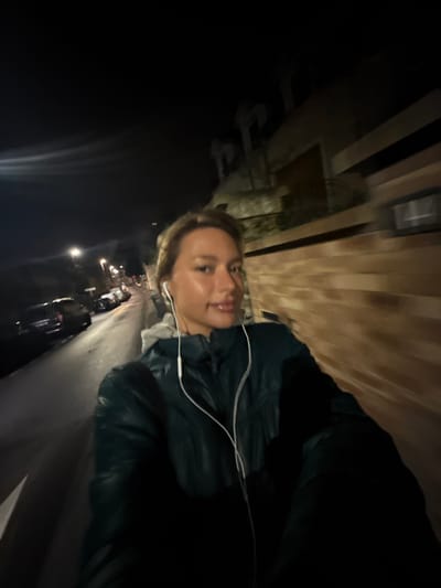 A woman with short blonde hair wears wired headphones. She's wearing a dark jacket, possibly leather, over a grey hooded top. She's on a street at night, with buildings visible behind her, some with wood siding. Cars are parked along the street. The lighting is dim, and the photo is taken at an angle.
