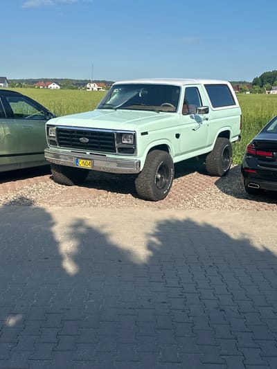 A light teal and white Ford Bronco SUV is parked on a gravel surface next to a black sedan. The Ford Bronco has large, black, all-terrain tires and a yellow license plate that reads "C-N04". The vehicle is facing slightly to the right. In the background, a field of green crops extends towards a line of houses with red roofs and a clear blue sky. The scene is illuminated by sunlight, casting shadows on the paved ground in the foreground. The location is identified as Czarnowo, Poland.