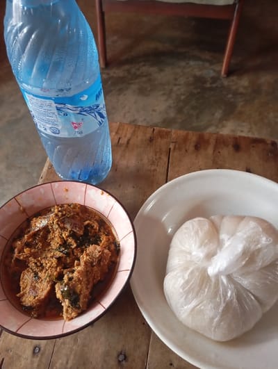 The scene depicts a meal laid out on a rustic wooden table in an indoor setting within Okere-Urhobo, Nigeria. A blue plastic bottle of "MP" branded water, marked "75cl," stands upright on the upper left, exhibiting condensation on its exterior. To the lower left, a pink-rimmed bowl contains a reddish-brown stew with visible pieces of meat or fish and dark green leafy vegetables. Adjacent to it on the lower right, a white ceramic plate holds a portion of white, starchy food, resembling fufu or eba, securely wrapped in a clear plastic bag. The wooden table surface displays a rough texture. The background is blurred, revealing a dark floor, possibly concrete, and the lower legs of what appears to be a wooden chair. No individuals are present in the image.