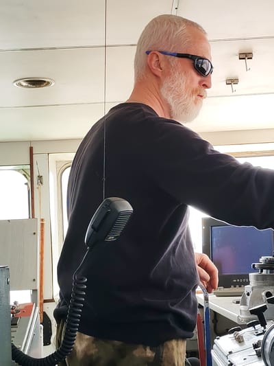 A man stands in what appears to be the bridge of a ship. He is white, with short white hair and a full white beard. He wears dark sunglasses with blue accents and a dark long-sleeved shirt. A microphone is attached to the console in front of him, its coiled cord extending down. A computer monitor and other electronic equipment are visible on the console. The interior is white with recessed lighting and hooks on the ceiling. The lower part of his body is obscured, but he appears to be wearing camouflage pants. The image shows no indicators regarding its location in the city of Koprino, Russia.