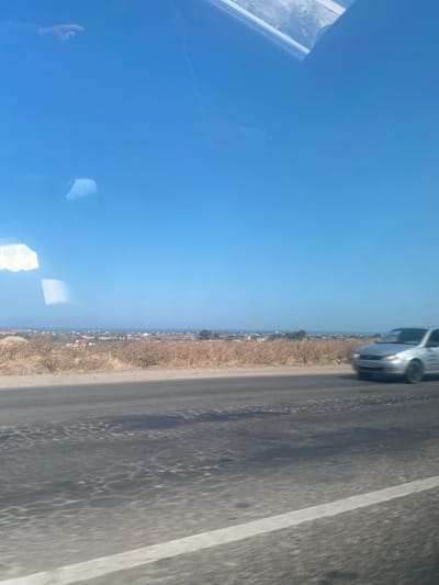 The image shows a bright blue sky with some reflections on what appears to be a car windshield. Below the sky, a hazy horizon line separates the sky from a landscape. The landscape features dry, brown vegetation in the foreground, transitioning to a scattered settlement of buildings in the middle ground. Further in the distance, a body of water, possibly the sea or a large lake, is visible. A silver car is driving on a dark asphalt road in the middle right of the image, moving from left to right. The road surface shows signs of wear and tear, with patches of uneven texture. The overall impression is of a sunny day in a somewhat arid coastal or lakeside area. There is no visible indication of the city of Dzhalgan, Russia, within the image.