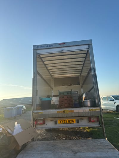The image displays the rear of a white box truck (lorry) with its hydraulic lift gate lowered to ground level. The truck's UK registration plate, YX22 WXG, is centrally visible on the rear bumper. The company name "JCPAYNE.COM" is displayed on the upper rear frame.
Inside the truck's open cargo bay, various items are stored, including a blue and black plastic bucket, several stacked red and green plastic crates (possibly for bottles or glassware), and a large, domed metal catering item, likely a chafing dish or serving pot.
In the immediate foreground, on the lowered lift gate, a partially open cardboard box contains white plastic bags and other discarded materials, with several black refuse bags positioned to its left. The ground beneath and around the truck consists of a mix of gravel and sparse green grass.
To the left of the truck, in the mid-ground, multiple blue wheeled bins are visible alongside a dark-colored car parked near a large, low-rise building with a dark roof, suggestive of an agricultural or industrial structure. To the right of the truck, a white pickup truck is parked on a grassy area.
The scene is illuminated by low-angle sunlight under a clear blue sky, indicating either early morning or late afternoon. The environment suggests an outdoor, possibly semi-rural or industrial setting within the city of Northallerton, United Kingdom.