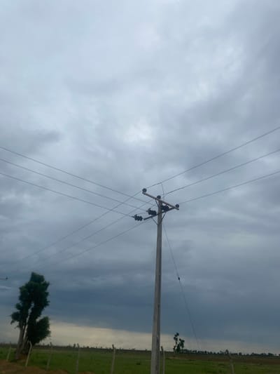 This media file captures a low-angle view of a wooden utility pole with multiple power lines strung across a cloudy sky. The pole is positioned in the lower right quadrant of the frame and is angled slightly upwards. Attached to the pole are several dark, spherical insulators. The power lines stretch diagonally across the image, disappearing into the overcast sky.
The scene appears to be an outdoor, rural setting. To the right of the utility pole, a strip of green, grassy land is visible, hinting at a field or pasture. In the distance, a line of trees or foliage marks the horizon, and faint signs of structures or fences can be discerned. A lone tree with dense foliage is present in the lower right portion of the image, contrasting with the sparse vegetation in the background.
The weather is overcast, with heavy, grey clouds dominating the sky, suggesting a possibility of rain or a generally gloomy day. The lighting is diffused due to the cloud cover, creating a soft, shadowless environment. The time of day is not definitively clear but is likely during daylight hours, given the overall illumination. There are no visible people or text in the image. The overall mood conveyed by the image is one of subdued natural beauty, with the stark lines of the utility pole and power lines creating a man-made element against the backdrop of a cloudy sky and rural landscape. The location context is Bulamare, Nigeria.