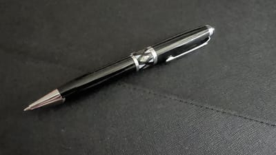 A black ballpoint pen with a silver clip and silver tip rests on a textured black surface. The pen has a decorative band around its barrel featuring a geometric pattern of alternating white and dark, possibly mother-of-pearl, inlay. The pen is positioned diagonally, with the tip pointing towards the lower left corner of the frame and the cap towards the upper right. The black surface appears to be a mat or a notebook cover with a subtle woven texture. A faint, diagonal seam or crease is visible on the surface to the right of the pen. The lighting suggests an indoor setting, likely a desk or table, with a soft, even illumination. No people or specific location cues are apparent beyond the provided context of Knittelfeld, Austria.