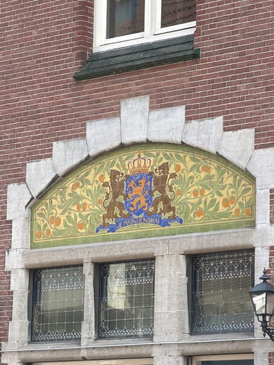 The image shows a detailed view of the facade of a brick building in Roermond, Netherlands. The building features a decorative tiled archway above a set of windows. The archway is adorned with a coat of arms depicting two lions holding a shield with a crown, surrounded by an orange and green floral pattern. Below the coat of arms, the inscription "JE MAINTIENDRAI" is visible. The windows below are framed by stone and feature decorative leaded glass. A streetlamp is visible on the right side of the image. The overall impression is one of architectural detail and historical significance.