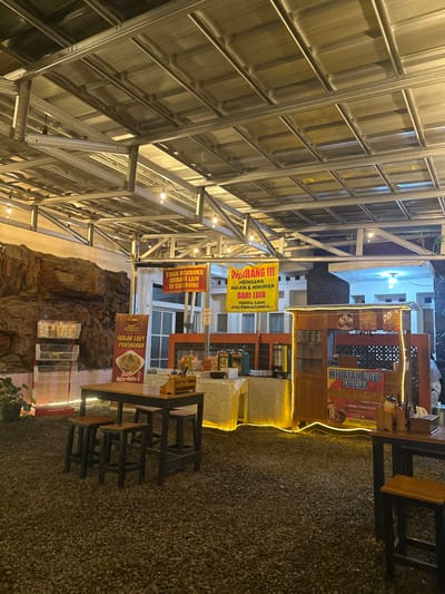 This image captures a semi-outdoor dining area at night, likely a food court or street food establishment, located in Kecamatan Cisaat, Indonesia. The ground is covered in gravel, and overhead, there's a large metallic roof structure supported by silver frames, fitted with numerous small lights illuminating the space.
Several food stalls or carts are visible, designed with wooden exteriors and decorated with warm yellow string lights. One prominent stall advertises "MIE AYAM COET SPECIAL" (Special Coet Chicken Noodles) and displays a phone number, "0896-7744-312". Another stall or sign promotes "SEBLAK COET PRASHANAN" (Prashanan Coet Seblak). Dark, square-shaped tables and matching stools are arranged on the gravel, indicating a casual seating area for customers.
Two yellow banners hang from the overhead structure. The larger one reads "DILARANG !!! MEMBAWA MAKAN & MINUMAN DARI LUAR TERIMA KASIH ATAS PERHATIANNYA" (FORBIDDEN!!! TO BRING FOOD & DRINKS FROM OUTSIDE THANK YOU FOR YOUR ATTENTION), indicating a policy of the establishment. The smaller banner states "TIDAK MEMBUKA CABANG LAIN DI SUKABUMI" (DOES NOT OPEN OTHER BRANCHES IN SUKABUMI). In the background, what appears to be a building with windows and some rock-like decorative elements are visible. The artificial lighting suggests it is evening or nighttime. No people are visible, but the setup implies a place for dining and social gathering.