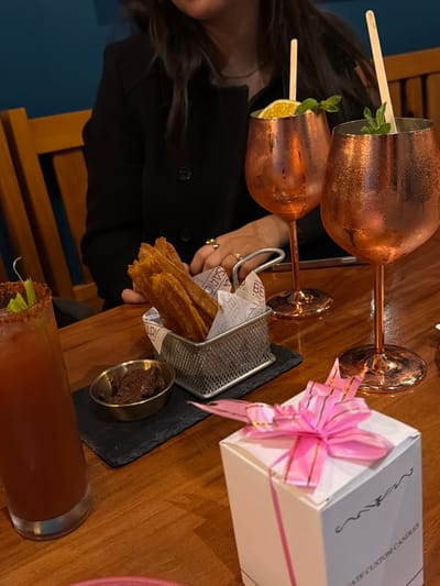 Two copper mugs with garnishes sit on a wooden table next to a white gift box with a pink bow. A black serving slate holds a metal basket filled with churros and a small dish of chocolate sauce. A bloody mary with a celery stalk garnish is also visible on the table, along with a person wearing a black top. The arrangement suggests a celebratory or indulgent occasion.
