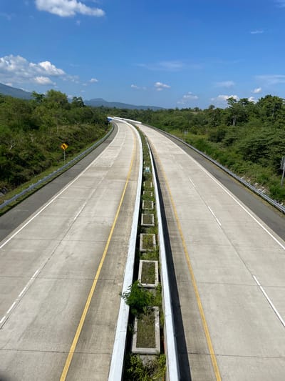 The image depicts a divided highway in Seulimeum, Indonesia, under a clear blue sky with scattered white clouds. The highway is made of concrete and has yellow and white lane markings. On the left side of the highway, there is a yellow road sign indicating a curve. The highway is flanked by lush green trees and vegetation on both sides, with hills or mountains in the distance. The central median of the highway features a planter box with greenery. The perspective is from an elevated position looking down the highway as it curves gently into the distance. There are no people or vehicles visible in the image.