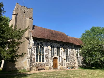 The image depicts St Peter's Church in Brome, Mid Suffolk, United Kingdom, a stone building with a tall tower and a tiled roof. The church has multiple arched windows, and a wooden bench sits in front of it on a lawn of dry grass. A wooden door is set in the middle of the front wall of the church. Trees partially obscure the building on both sides, and the sky is clear and blue.