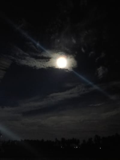 The image depicts a night sky with a full moon partially obscured by clouds. A bright beam of light emanates from the moon, cutting diagonally across the frame. Silhouetted trees form a dark line across the bottom of the image, suggesting a rural or natural landscape at the edge of a town. There are a few faint lights visible amongst the trees, possibly from buildings or distant streetlights. The overall impression is one of a serene and atmospheric night scene.