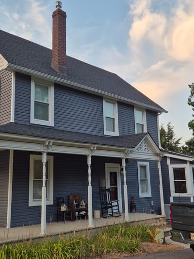 The image shows a two-story house with dark blue siding and a dark gray roof. A prominent brick chimney with a metal cap extends from the roof. The house features a wrap-around porch with white columns and decorative trim. Several windows with white trim are visible on both floors. On the porch, there are a rocking chair, various decorative items, and some outdoor furniture. In front of the house, there is overgrown grass and a rusty metal watering can with decorative flowers. A portion of a dark green pickup truck is visible on the right side of the image. The sky above is a mix of blue and light peach-colored clouds. The image appears to have been taken from a slightly low angle, looking up at the house. The overall scene suggests a residential property.