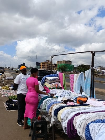 A man in a white t-shirt and orange hat stands next to a woman in a pink dress. They are browsing a sidewalk vendor's display of folded and draped textiles, including towels and sheets. A black plastic stool holds a cup. Bags of produce are nearby. The scene is alongside a road with highway lighting and buildings visible in the background under a cloudy sky.