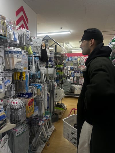 An indoor scene within a retail store in Shinagawa, Japan, shows a male individual in profile, facing right, actively browsing merchandise. He is wearing a black beanie, glasses, a dark hooded jacket, and a dark overcoat. His right hand is not visible. A white plastic shopping basket with pink handles is positioned at his lower right. The store features tall shelving units densely packed with various household goods, predominantly cleaning supplies, kitchen tools, and utility items, many packaged in clear plastic with Japanese labeling. These items include brushes, sponges, mops, and squeegees. A prominent red arrow graphic points upwards on a white wall section above the merchandise. The store has light-colored floor tiles, a suspended ceiling with visible ceiling tiles, and multiple fluorescent strip lights providing illumination. Additional aisles with similar merchandise displays are visible in the background, further into the store.
