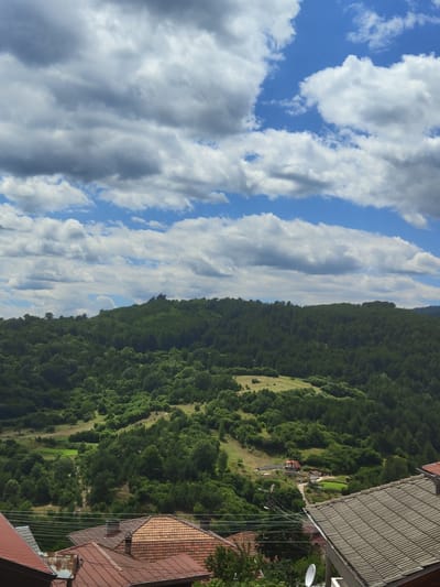 This is a high-angle, medium shot photograph taken in Pletena, Bulgaria. The image captures a landscape dominated by rolling hills covered in dense green forest. A small, rural settlement is visible nestled within the trees, characterized by a few red-roofed houses and a satellite dish. In the foreground, the tiled roofs of several houses are prominent, with power lines running horizontally across the frame. The sky above is a vibrant blue, dotted with numerous white and grey cumulus clouds. The overall scene depicts a tranquil, natural environment with evidence of human habitation.