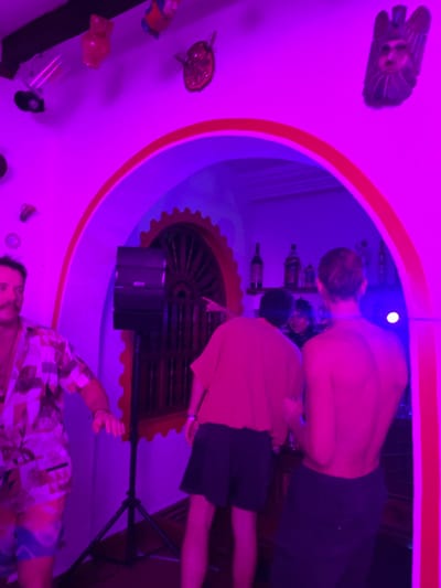 This image captures an indoor social gathering under intense purple ambient lighting in Fortín de las Flores, Mexico. The foreground features a white wall adorned with various decorative objects, including pottery pieces and carved wooden masks, illuminated by a spotlight from the upper left. A prominent archway, trimmed in red and white, frames the central part of the scene, leading into an adjacent room or area.
To the left of the archway, a male subject with a mustache, wearing a patterned short-sleeved shirt and patterned shorts, is partially visible, appearing to be in motion. A black audio speaker on a tripod stand is positioned near the left side of the arch.
Through the archway, at least three additional individuals are visible. In the center, a male subject, viewed from behind, wears a light-colored t-shirt and dark shorts; he is pointing his right index finger towards the right side of the frame. To his right, another male subject, also viewed from behind, is bare-chested and wears dark pants, with his hair tied back. Both appear to be moving within the space. Deeper within the archway, behind the pointing subject, a fourth individual is partially visible, illuminated by a bright light source, and appears to be smiling.
The background visible through the archway includes a bar or shelf displaying several bottles. A large, circular wooden decorative element, resembling a spoked wheel or ornate grating, is fixed to the wall on the left side of the inner archway.