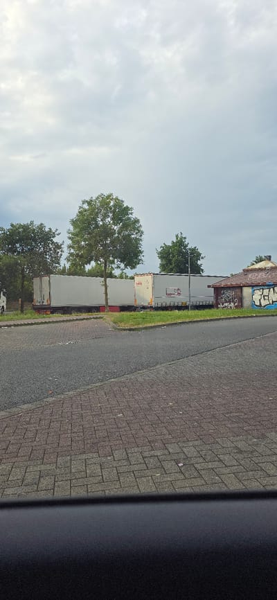 A row of parked semi-trailer trucks is visible on the left side of the image, facing away from the viewer. The trucks are white and appear to be identical in model. Some have logos or writing on their sides, but they are not clearly legible. The trucks are parked on a paved surface next to a grassy area. Behind the trucks, trees and a building with graffiti are visible. The building appears to be an industrial or warehouse structure. The sky is mostly clear with some light clouds. The image is taken from a low angle, possibly from a vehicle. The time of day appears to be daytime, and the weather seems to be mild. The scene is set in an industrial or commercial area, possibly a parking lot or loading dock. There are no people visible in the image. The image appears to be taken in Staufenberg, Germany.