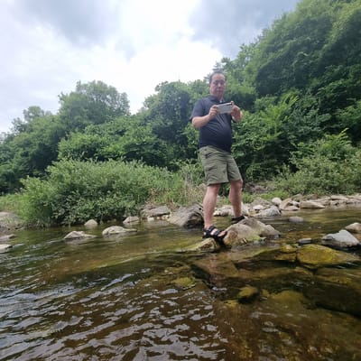 A man is standing on rocks in a shallow, clear river. He is wearing a black polo shirt and khaki shorts, and black sandals. He is holding a smartphone in his hands, as if taking a picture or recording a video. The riverbed is visible through the water and is covered with rocks and pebbles. Lush green trees and bushes line the banks of the river, creating a natural, serene environment. The sky is overcast with gray clouds. The scene appears to be in a natural outdoor setting, possibly a park or a hiking trail. The location of Mureungdowon-myeon, South Korea, suggests a rural or mountainous area with natural beauty, consistent with the image.