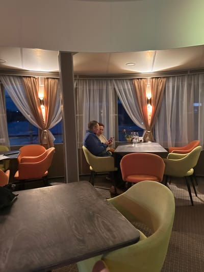 The image depicts an indoor restaurant seating area with a curved window offering a view of the exterior. Two individuals are seated at a table with glasses and a small plant. The restaurant features orange and yellow upholstered chairs, a dark rectangular table in the foreground, and sheer white and brown curtains. The lighting suggests it is evening. The image does not contain any information to suggest it is located in the city of Doksha, Russia.