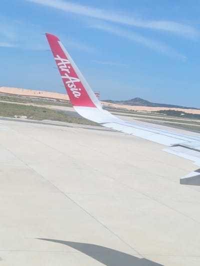 The image shows the wing of an AirAsia airplane on the tarmac at Cam Ranh International Airport (CXR) in Cam Ranh City, Vietnam. The wing is predominantly white with a red tip featuring the "AirAsia" logo in white lettering. The airplane appears to be stationary on a concrete runway or taxiway, with clear lines and segmented concrete panels visible. In the background, there are sand dunes and sparse vegetation, characteristic of the coastal environment of Cam Ranh. A control tower is partially visible in the distance. The sky is blue with some wispy white clouds. The lighting suggests it is daytime. There are no people or other vehicles visible in the immediate vicinity of the wing. The shadow of the airplane wing is cast on the tarmac.