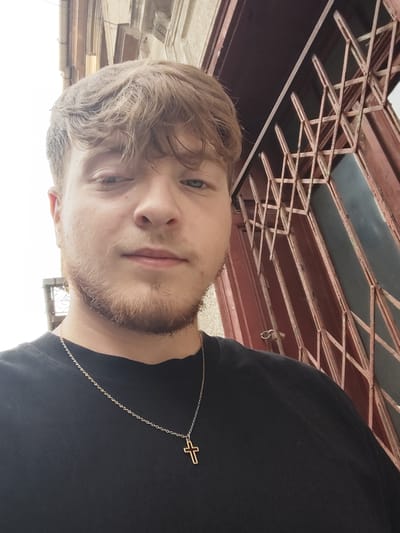 A close-up, waist-up selfie of a young white male with light brown, tousled hair and a light beard. He is looking at the camera with a neutral expression, his blue eyes clearly visible. He is wearing a black t-shirt and a silver chain with a small gold cross pendant. The background appears to be an urban setting in Arad, Romania, with a weathered, reddish-brown metal gate or window grate to his right, and a building with light-colored stonework above and to the left. The lighting suggests it is daytime, possibly overcast.