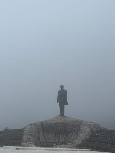 A statue of a man in a suit holding a briefcase stands on a rock formation. The statue is surrounded by mist, obscuring the background. The rock formation is part of a larger structure that appears to be a monument or memorial. There are tiered flowerbeds surrounding the base of the rock formation, with some flowers visible. The overall scene suggests a somber or contemplative atmosphere due to the fog and the subject matter. The location is specified as the city of Chang Khoeng, Thailand.