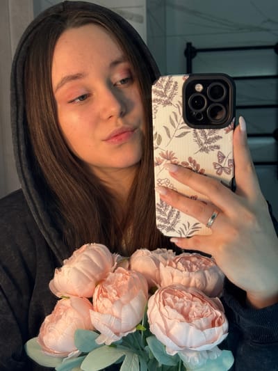 The image depicts a female individual taking a mirror selfie in an indoor setting within Prikubanskii, Russia. The environment appears to be a bathroom, characterized by a light-colored tiled wall with a subtle pattern and a dark-colored, possibly metallic, heated towel rail visible in the reflection.

The female has long, dark brown hair, partially obscured by a dark grey hooded sweatshirt or jacket. She has light-colored skin and is wearing minimal makeup, including a pink eyeshadow and a gloss on her lips. She holds a smartphone in her right hand, which is equipped with a patterned case featuring a light background with floral designs (leaves, small flowers) and butterflies. The phone's multi-lens camera array is prominent. Her fingernails are manicured with a light polish, and a silver-colored ring is visible on the fourth finger of her right hand. Held in her left arm, partly visible, is a bouquet of approximately seven to eight large, light pink peonies with green leaves. The lighting is warm, highlighting her face and the flowers.
