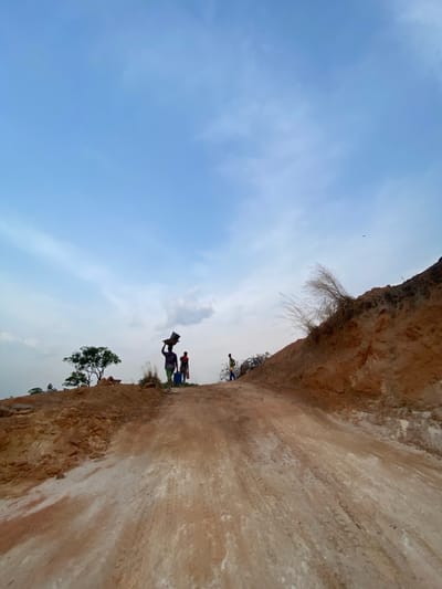 The image captures an outdoor scene in Asaga Amangwu, Nigeria, centered on an ascending dirt road. The road surface is composed of reddish-brown soil with significant patches of lighter, possibly whitish, earth. To the right, a steep embankment of reddish-brown earth is visible, topped with dry, light-colored grasses. To the left, the terrain is also earthen and rugged. Three individuals are present on the road. In the mid-ground, a person wearing dark clothing carries a dark, rectangular basin or container balanced on their head. Immediately behind this individual, another person in dark attire stands near a blue container on the ground. Further up the path to the right, a third individual, clad in lighter-colored clothing, walks away from the viewer. A small tree with green foliage is discernible on the horizon to the far left. The sky is largely clear and light blue, with sparse, wispy clouds. A small, dark, winged object is visible in the upper right quadrant of the sky.