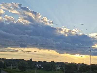 A landscape photograph captures a dramatic sunset over a rural scene in Ateptsevo, Russia. The sky is a prominent feature, dominated by a thick band of dark, heavy clouds stretching diagonally across the frame from the upper left to the lower right. Streaks of sunlight pierce through the clouds, creating crepuscular rays that fan out into the clear blue sky above. The lower part of the sky, near the horizon, is ablaze with the warm, golden hues of the setting sun, illuminating the undersides of the clouds with a bright yellow and orange glow.
Below the sky lies a silhouette of a treeline, dense and dark, suggesting a wooded area. Scattered within this treeline and on the visible fields are a few houses, one with a distinct white roof and a few smaller structures. Power lines and utility poles are visible in the mid-ground, crossing the scene horizontally and vertically, indicating human presence and infrastructure in this rural setting. The foreground is a blur of green foliage, suggesting the photograph was taken from a moving vehicle or a slightly elevated position overlooking the landscape.
The time of day is clearly sunset, given the vibrant colors and the angle of the sun. The weather appears to be dynamic, with both stormy-looking clouds and clear sky present, creating a visually striking contrast. The overall mood evoked is one of natural beauty and the calm transition from day to night, with a touch of the dramatic provided by the cloud formations and the piercing sunlight. There is no visible text in the image.