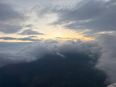 This media file presents an aerial view from an airplane, capturing a dramatic sky and cloudscape during either sunrise or sunset. The upper portion of the image reveals a vast sky dominated by scattered, varied clouds ranging from light gray to white. Warm, golden-yellow light diffuses through gaps in the clouds along the horizon line, casting a soft glow that contrasts with the darker, higher cloud formations.
Mid-frame, a dense and expansive layer of white, fluffy cumulus clouds stretches horizontally, appearing thick and substantial. Below this thick cloud cover, the ground is visible but largely obscured and darkened by shadows and distance. Faint outlines of land features, possibly agricultural fields or forests, can be discerned. A thin, light-colored line, likely a road or highway, winds across the dark landscape, with a few distant, small clusters of lights or structures suggesting human habitation. A small part of the aircraft's wing or engine nacelle is visible in the bottom right corner, confirming the aerial perspective.
The scene conveys a sense of peaceful grandeur and the immense scale of the sky and landscape from above. The time of day is clearly marked by the warm, low-angle light on the horizon, indicating dawn or dusk. The weather is characterized by extensive cloud cover at multiple altitudes. No visible text is present in the image. Based on the provided context, the image likely depicts the area around Vecsés, Hungary.