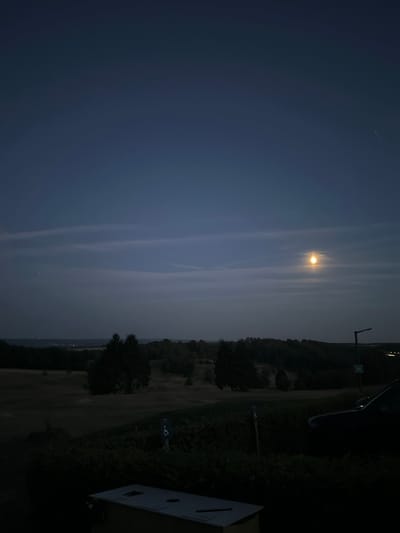 An outdoor, elevated night scene in Méan, Belgium features a dark sky with a prominent full moon glowing brightly in the upper right quadrant. Wispy clouds streak across the sky. Below the sky, a dark, rolling landscape is visible, characterized by a treeline and what appears to be a golf course or open field with scattered trees. In the foreground, a dark car is parked on the right side. A lamppost stands near the car. On the left side of the foreground, a light-colored waste receptacle or structure is partially visible. The overall atmosphere is tranquil and dimly lit by the moon.