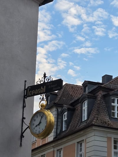 The image shows a vintage golden clock sign hanging from a building in Ansbach, Germany. The sign reads "Tornahl Uhren Schmuck Juwelen," indicating it is for a watch, jewelry, and gem store. The clock itself has Roman numerals and a visible time of approximately 8:10. The building behind the sign appears to be old, with a tiled roof and arched windows. The sky is blue with scattered white clouds. The lighting suggests it is daytime, likely late afternoon or early evening. There are no people visible in the image.