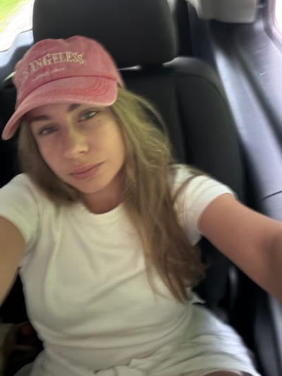 A young Caucasian female is seated in the passenger seat of a vehicle. She is wearing a pink baseball cap with "Los Angeles" embroidered on it and a white t-shirt. The female is looking towards the camera with a neutral expression. Her blonde hair is visible beneath the cap and falls over her shoulders. The background shows the interior of the car, with black leather seats and part of the headrest. The lighting suggests it is daytime, possibly with sunlight filtering in from outside. No specific location in Žunovnica, Bosnia and Herzegovina can be determined from the image.
