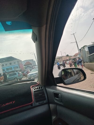 The image captures a vibrant daytime street scene in Akobo, Nigeria, viewed from inside a vehicle. The perspective looks out the right side of the car, encompassing views through the side window and reflected in the exterior side mirror.
The car's interior elements are visible, including a portion of the dashboard with an air vent, a door panel with a handle, and the car's roof lining. Prominently featured on the dashboard is a black mat with a red border, which clearly displays the word "CAMRY" in red lettering, indicating the make of the vehicle.
Outside the car, the street is bustling with activity. Numerous people are visible, many riding motorbikes, which are common modes of transport. Other vehicles, including cars and vans, are also present, either parked or in motion. Buildings line the street, suggesting an urban or semi-urban environment. Overhead, utility poles and a network of wires crisscross against a bright, clear sky, indicating fair weather conditions. The overall scene depicts a typical busy day on the streets of Akobo.