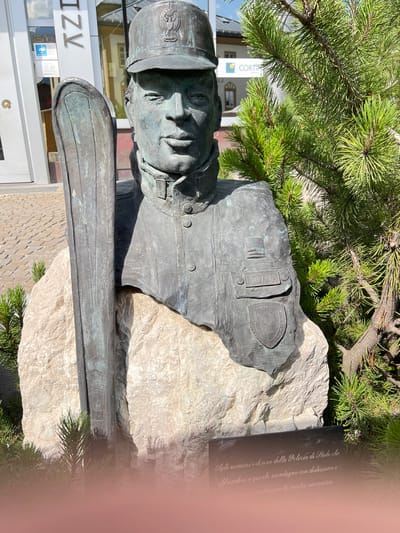 This is a photograph of a bronze bust of a man in a uniform, likely a police officer, given the inscription "POLIZIA" visible on his uniform. He is wearing a cap with an insignia on the front. The bust is mounted on a rough, light-colored stone.
Behind the bust, a pine tree branches out, its green needles providing a contrast to the bronze and stone. In the background, a building with windows is visible, with the partial word "IZV" and other text in Italian on the facade. The building appears to be modern, with glass doors.
A dark plaque with an inscription in Italian is affixed to the stone behind the bust. The visible text on the plaque reads: "Agli uomini e donne della Polizia di Stalo che offrendosi a queste montagne con dedizione e santiscono la nostra sicurezza." This translates to: "To the men and women of the State Police who, offering themselves to these mountains with dedication and our safety."
The scene appears to be outdoors, possibly in a town square or public space, given the building in the background. The sunlight suggests it is daytime, likely a clear day with no visible clouds. The overall impression is one of commemoration and respect for law enforcement.