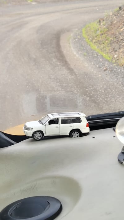 A miniature white SUV toy car is positioned on a textured, light-colored surface, possibly the dashboard of a vehicle. The car is facing left, with its wheels slightly turned. In the background, a blurred, dusty road curves to the right, bordered by rough terrain with scattered rocks and sparse green vegetation. The lighting suggests an outdoor, daytime setting. The specific location in Yerystivka, Ukraine, cannot be determined from the visual information provided.
