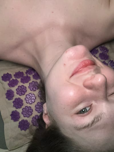 The image presents an overhead, close-up view of an individual, likely a young adult, lying on their back. The visible portion includes the person's upper chest, neck, and the right side of their face, which is slightly angled towards the camera. The skin tone is fair, with some areas of localized redness on the cheeks. The individual has light-colored eyes, appearing blue-green, and their right eye is partially visible and open. A faint smile or smirk is discernible, with the lips slightly parted and appearing moisturized. Dark hair is visible along the lower right edge of the frame. The person's head and neck rest on an acupressure mat, which is beige or light brown. This mat is patterned with numerous purple, rosette-shaped, spiky elements arranged in a grid. No identifiable environmental details are present to determine a specific location within Matiushino, Russia, or any other setting.