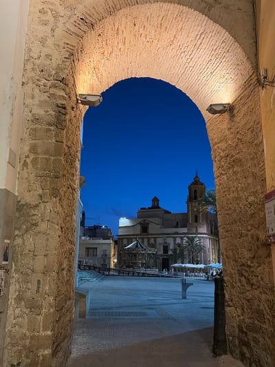 The image shows an arched stone passageway looking out onto a plaza in Cádiz, Spain. The sky is a deep twilight blue. In the distance, across the plaza, is a church with a prominent bell tower. The plaza itself appears to be a public space with a carousel, outdoor seating with umbrellas, and paved ground. Lights are visible illuminating the stonework of the archway and the plaza. The stone of the archway is weathered and textured. The overall impression is of an evening scene in an historic urban setting.