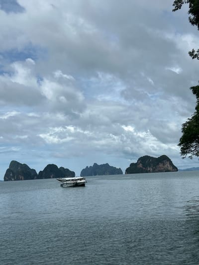 The image depicts a serene seascape with several limestone karsts rising from the water under a cloudy sky. A white boat with a covered section is anchored in the foreground, floating on the calm, blue-grey water. In the background, a cluster of karsts are visible, some appearing larger and more prominent than others, all covered in lush green vegetation. The sky is filled with a mix of light and dark grey clouds, suggesting an overcast or partly cloudy day. The overall impression is one of natural beauty and tranquility. While the specific location in Tambon Krasom, Thailand, is mentioned, the image itself does not contain any overt markers to definitively identify it as such, though the geographical features are consistent with the karst landscapes found in regions like Phang Nga Bay. There are no people or animals visible in the image.